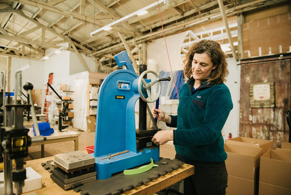 white woman wearing a dark green sweatshirt in a workshop in front of a piece of blue equipment appears to be making something
