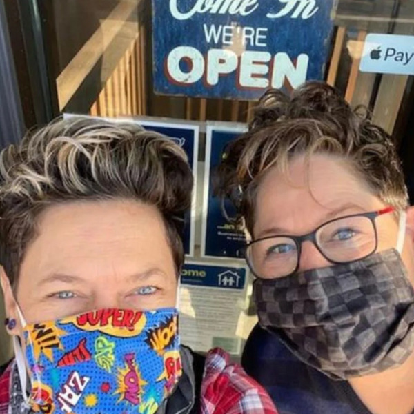 two white woman, one wearing glasses, both wearing masks, come in we're open sign in the background