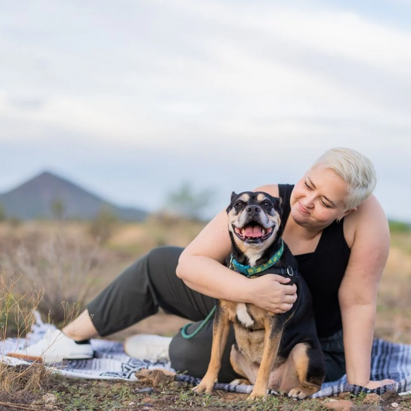 woman with short blonde hair, sitting in a field, leaning towards a black and brown down, almost hugging him, he is wearing a teal biothane long line