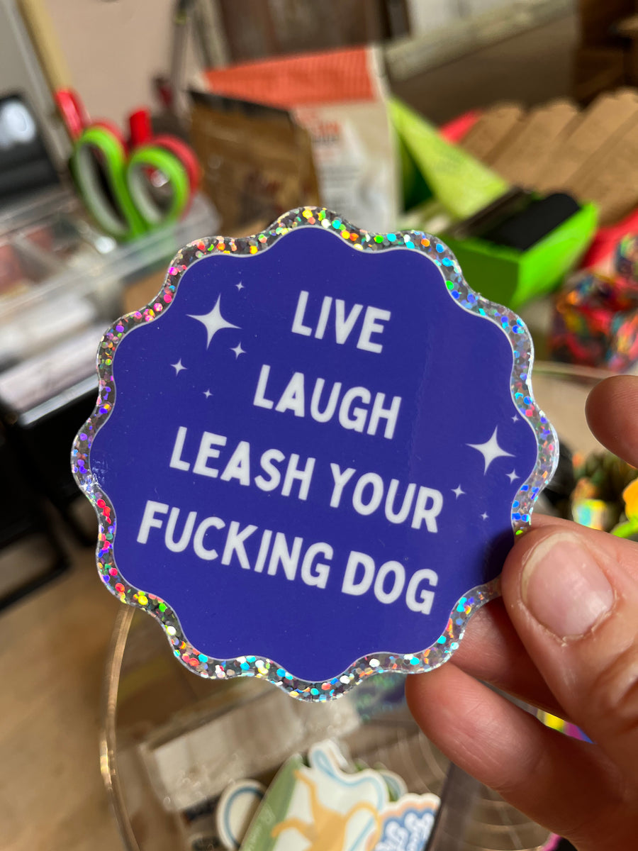 close up of person holding blue and silver sticker that says "Live Laugh Leash your $ucking Dog"