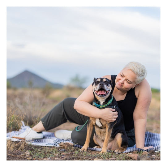 woman with short blonde hair, sitting in a field, leaning towards a black and brown down, almost hugging him, he is wearing a teal biothane long line