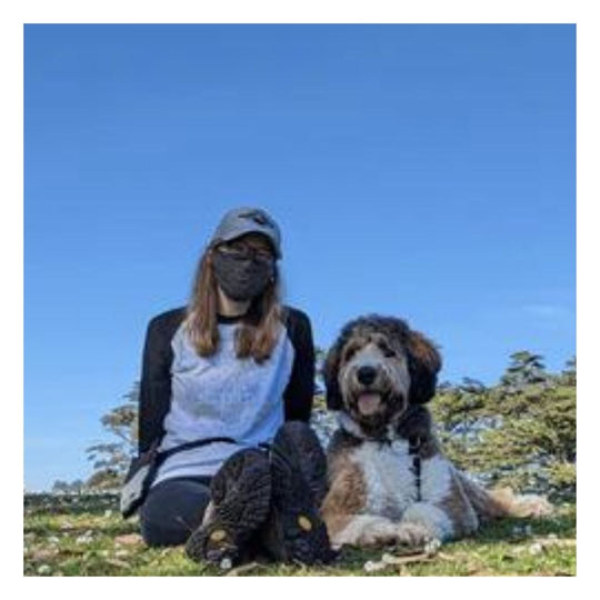 woman with long blonde hair wearing a grey hat, black mask, and white and black shirt, sitting in the grass next to a tan and white large curly haired dog. 