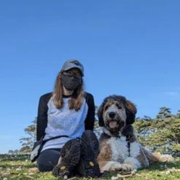 woman with long blonde hair wearing a grey hat, black mask, and white and black shirt, sitting in the grass next to a tan and white large curly haired dog. 