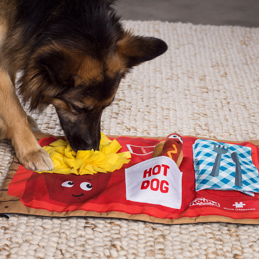 plush dog puzzle mat with french fries, hotdog and napkin on the front in red, yellow and blue with brown dog sniffing the french fries dog