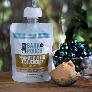 White, tan, and blue bark pouch in peanut butter & blueberry recipe on table with a scoop of peanut butter and bowl of blueberries