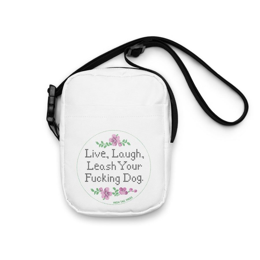 white crossbody bag laying flat with black strap reads in cross stitch font "Live, Laugh, Leash your F-ing Dog