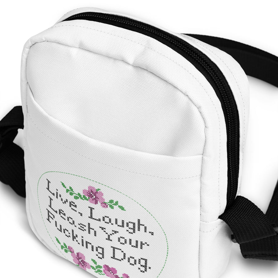 close up of outside front of white crossbody bag that reads in cross stitch font "Live, Laugh, Leash your F-ing Dog