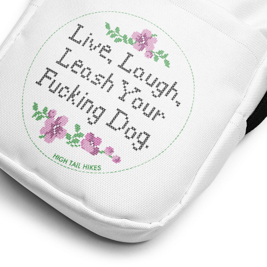 close up of white crossbody bag that reads in cross stitch font "Live, Laugh, Leash your F-ing Dog