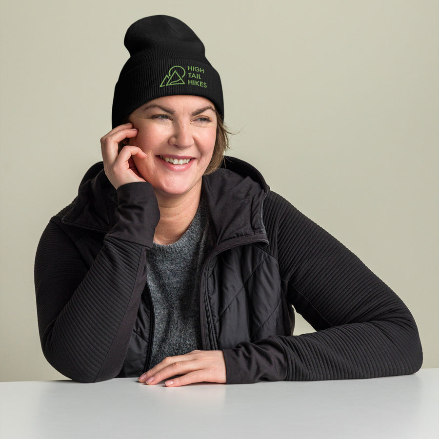 woman wearing a warm coat and black cuffed beanie with green high tail hikes logo looking to the right