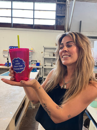 women smiling while holding a pink cup with green straw and blue and silver sparkle sticker that says "Live Laugh Leash your &ucking Dog"