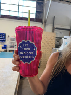 persons hand holding a pink cup with green straw and blue and silver sparkle sticker that says "Live Laugh Leash your &ucking Dog"