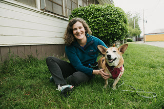 Founder Liz Williams sitting on grass with a dog wearing a red harness in front of a house.