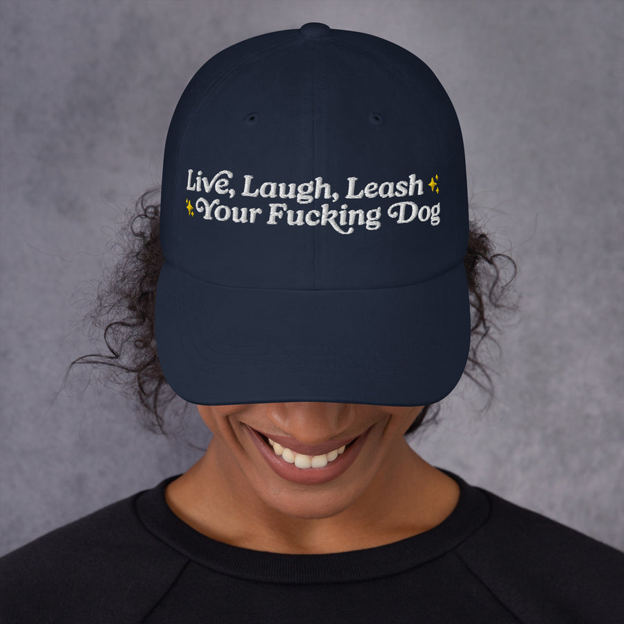 woman wearing dark blue dad hat with head down to show white letters that read "Live Laugh Leash Your F Dog"