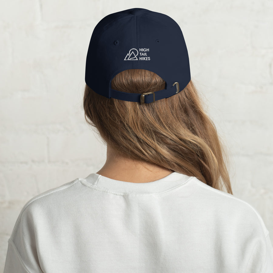 back of womans head, she is wearing a white sweater, and dark blue dad hat with white high tail hikes logo above the adjustable strap