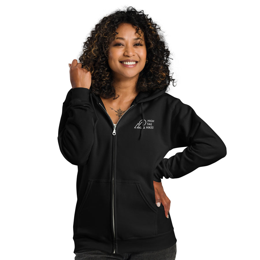 female with dark curly hair wearing black zip up hoodie - showing the front zipper and white high tail hikes logo in the front left chest area