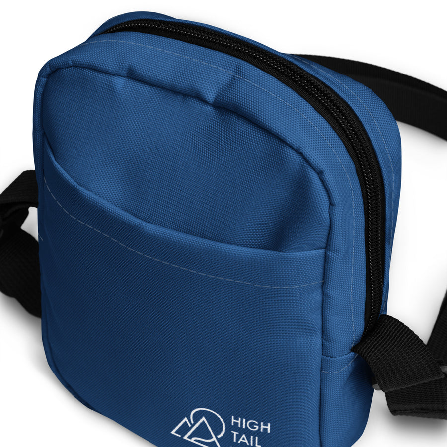 close up top view of blue crossbody bag, white high tail hikes logo on bottom of pocket