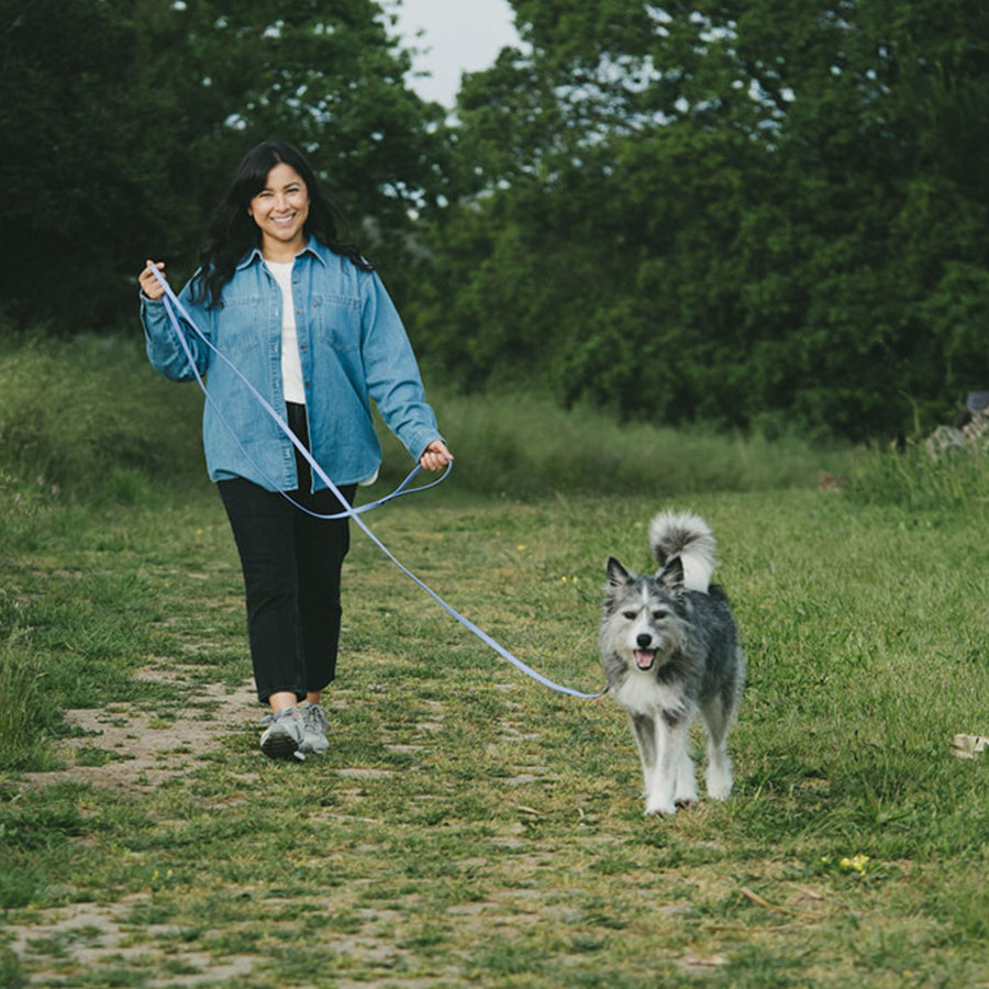 Woman walking a dog on a long biothane leash in a grassy field with trees in the background