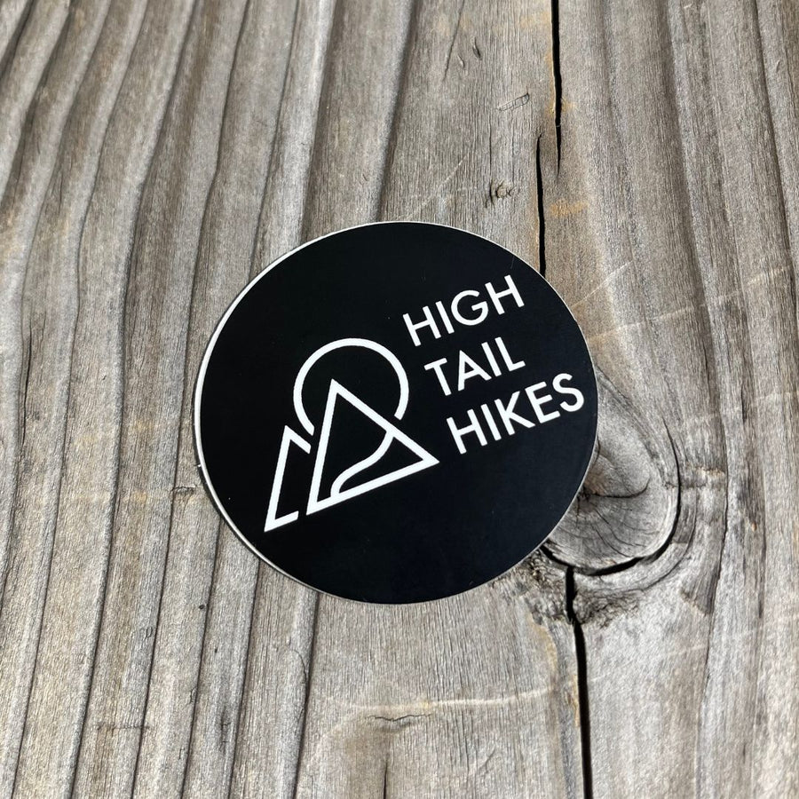 round black sticker with white High Tail Hikes logo on a wood background